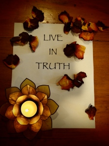 A sprinkling of rose petals frame the words Live in Truth with a small burning candle set inside a glass flower placed on the lower left corner.